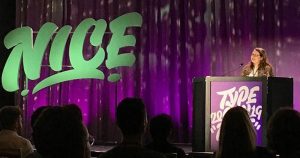 A purple backdrop on a stage, a small person standing behind a podium and the word "NICE" in teal on the stage next to her