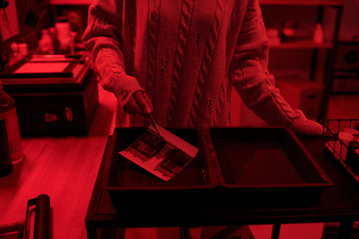 Person developing a photographic print in a darkroom, hands placing a photo into a chemical tray under red safety lighting.